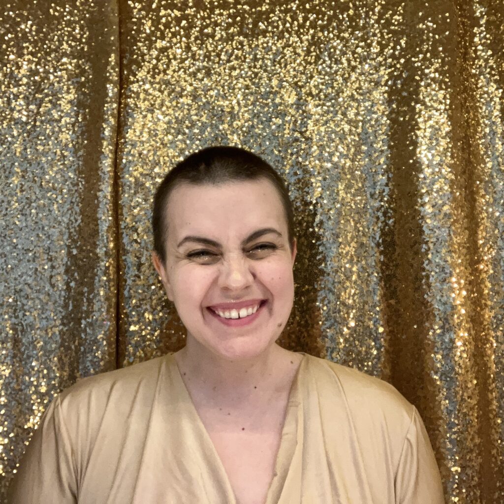 Photograph of Vero Rose Smith, a person with short dark har and pale skin smiling and standing center frame. The background is made of golden sequins.